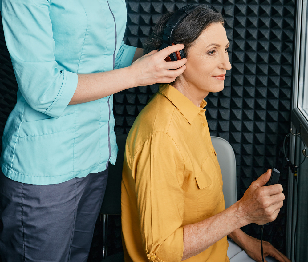 A woman in a yellow shirt sits in a soundproof room, wearing headphones, while a professional adjusts them. The woman holds a response button, likely undergoing a hearing test.