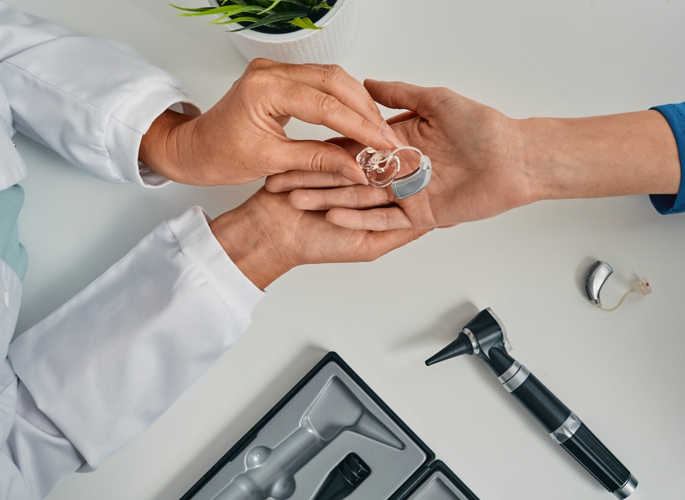 A doctor hands a hearing aid device to a patient. An otoscope, hearing aid accessories, and a potted plant are visible on the white table.