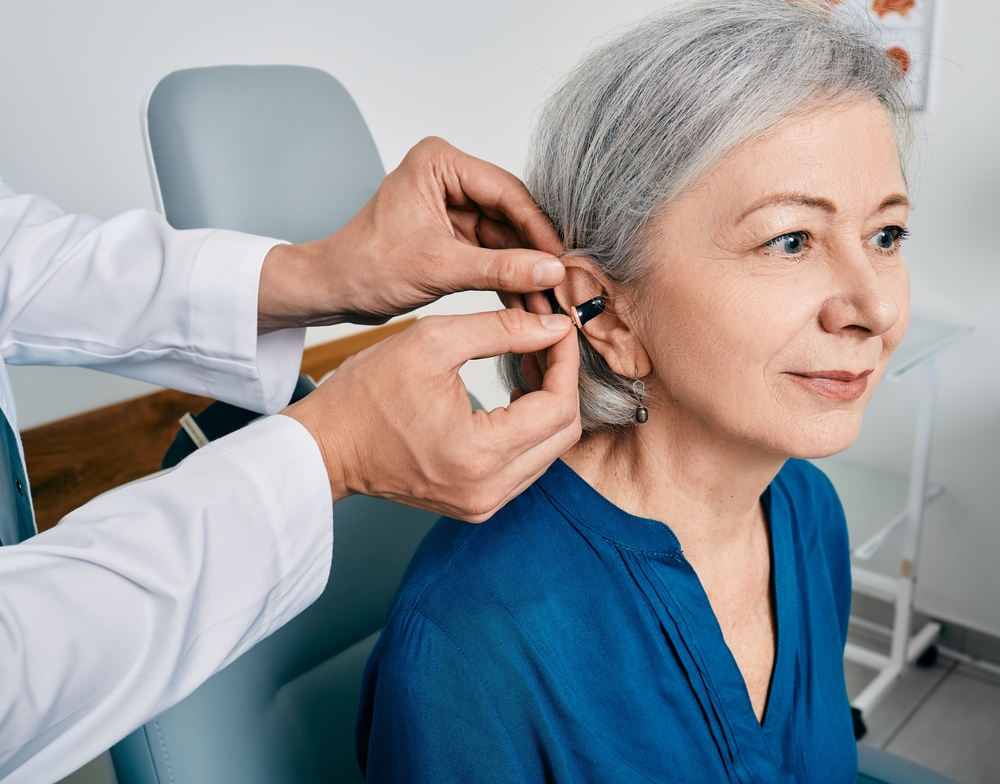 A healthcare professional fits a hearing aid behind the ear of an older woman with gray hair, who is seated and wearing a blue blouse, in a clinical setting.