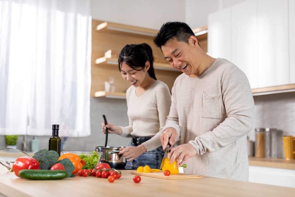 A man slices a yellow bell pepper while a woman stirs a pot on the stove. Fresh vegetables are on the kitchen counter. Both are smiling and cooking together in a bright, modern kitchen.