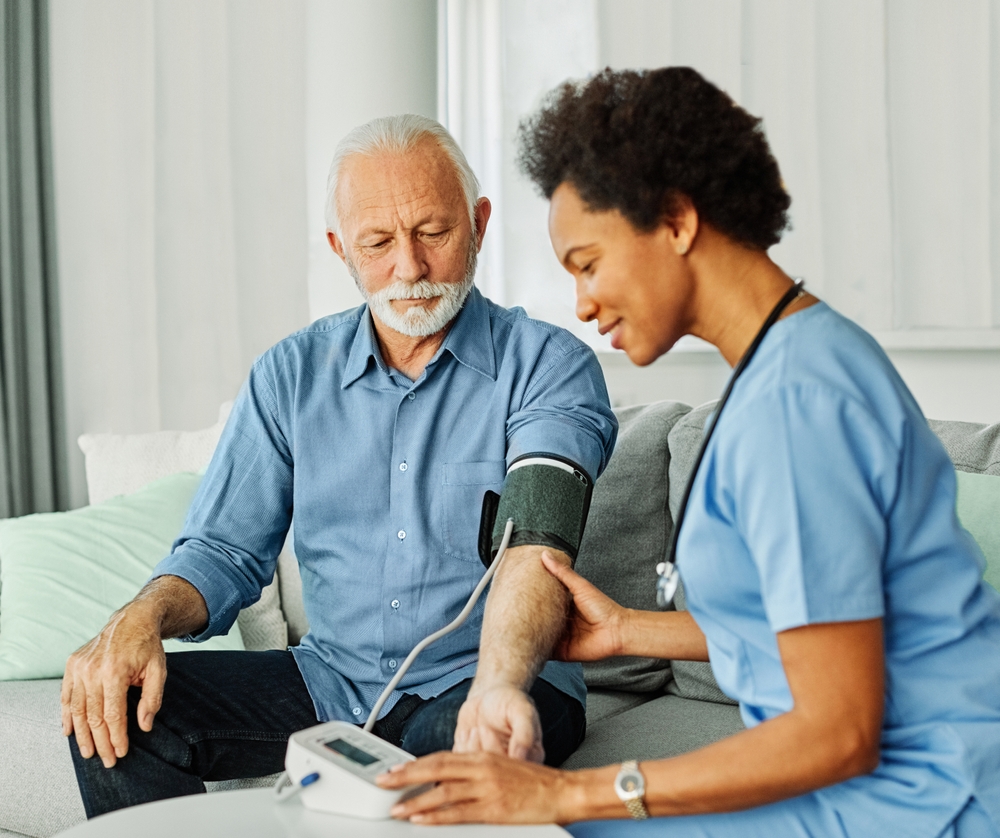 A nurse in blue scrubs measures the blood pressure of an older man with gray hair and a mustache, who is sitting on a couch with his sleeve rolled up. The nurse is checking the monitors reading.