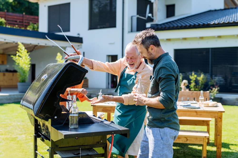 Two men stand outside by a barbecue grill, smiling and talking. One wears an apron and holds grilling tongs, the other stands beside him. Picnic tables and a modern house are visible in the background on a sunny day.