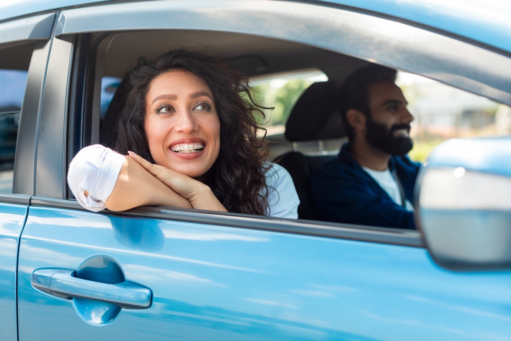 A young woman with curly hair and braces smiles while leaning out of a car window. A bearded man sits in the driver’s seat, both appearing happy and relaxed in the blue car.