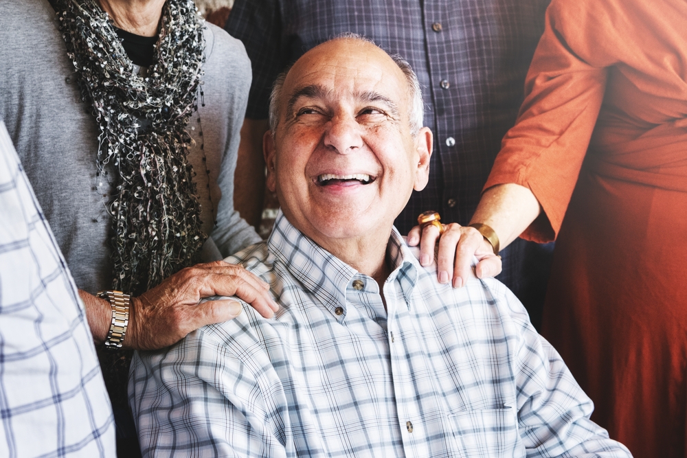 An older man in a plaid shirt smiles joyfully while surrounded by friends, who rest their hands supportively on his shoulders. The group appears happy and engaged in a warm, friendly moment.