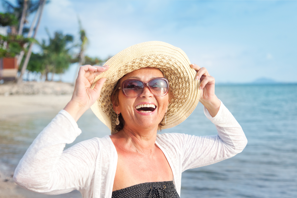 Smiling woman wearing sunglasses and a sun hat stands on a sandy beach near the ocean, with palm trees and blue sky in the background. She looks happy and is enjoying a sunny day.