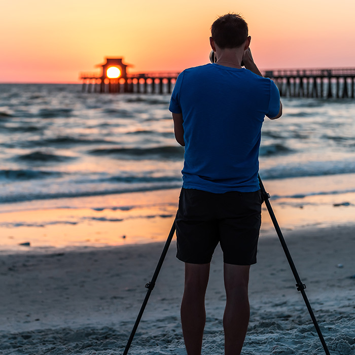 A person in a blue shirt and shorts, who experiences hearing loss, uses a camera on a tripod to photograph the sunset over the ocean, with a pier in the background.