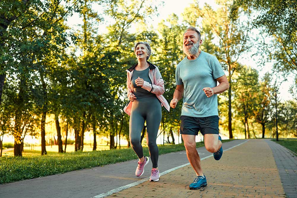 A smiling older couple jogs together on a paved path in a park, surrounded by green trees and sunlight. The woman wears athletic clothes and the man wears a t-shirt and shorts. Both appear happy and energetic.