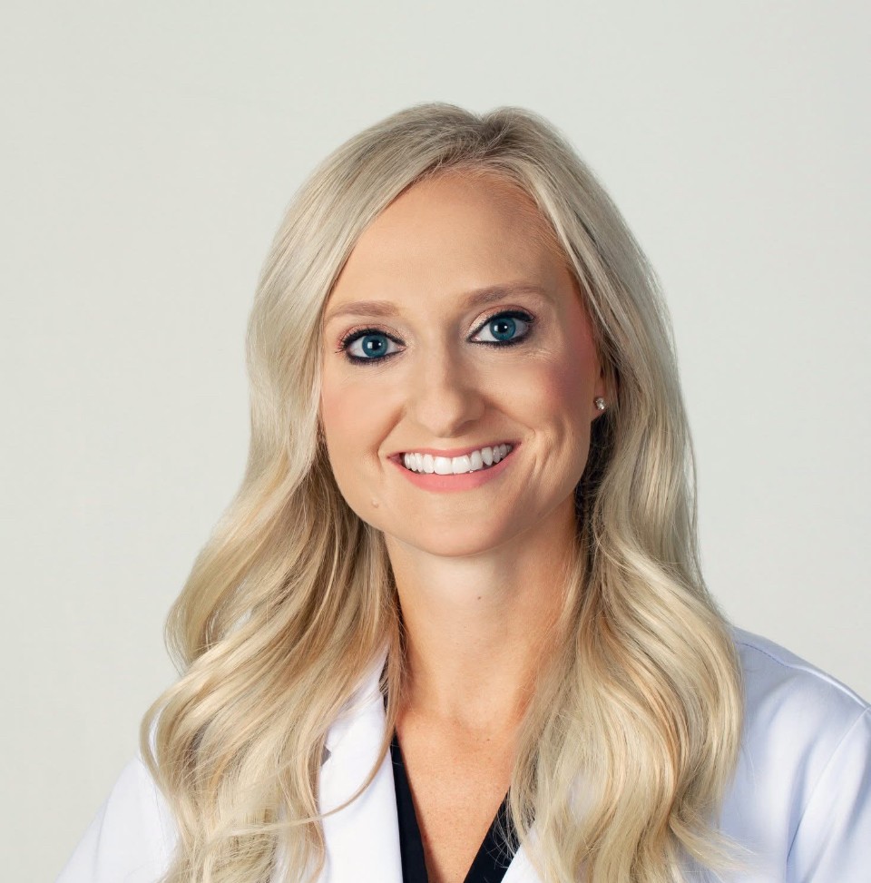 A smiling woman with long blonde hair wearing a white medical coat, posing against a plain light background.