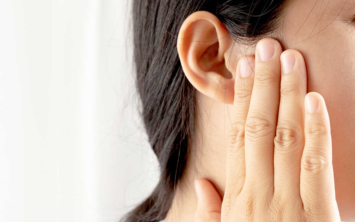 A person with dark hair holds their hand to their ear, covering it as if experiencing discomfort or pain, with a neutral background.
