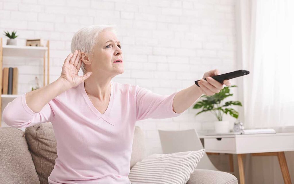 An older woman with short white hair sits on a couch holding a TV remote, raising her other hand to her ear as if trying to hear better. She looks focused in a bright, modern living room.