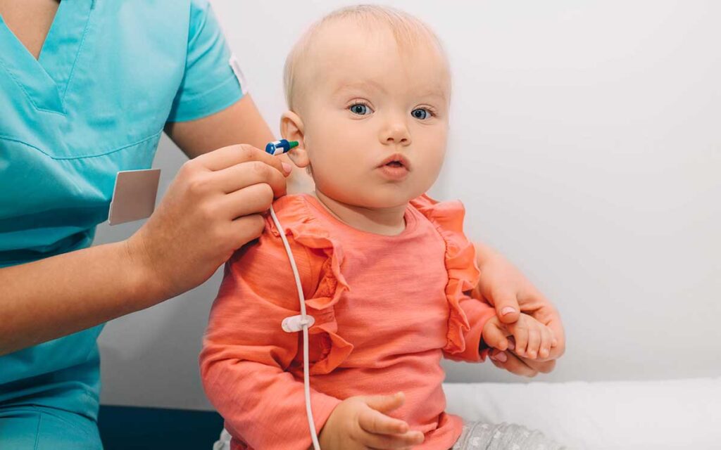 A healthcare professional tests a baby’s hearing by placing an ear probe in the babys ear. The baby, wearing a pink top, sits on an exam table and looks forward.