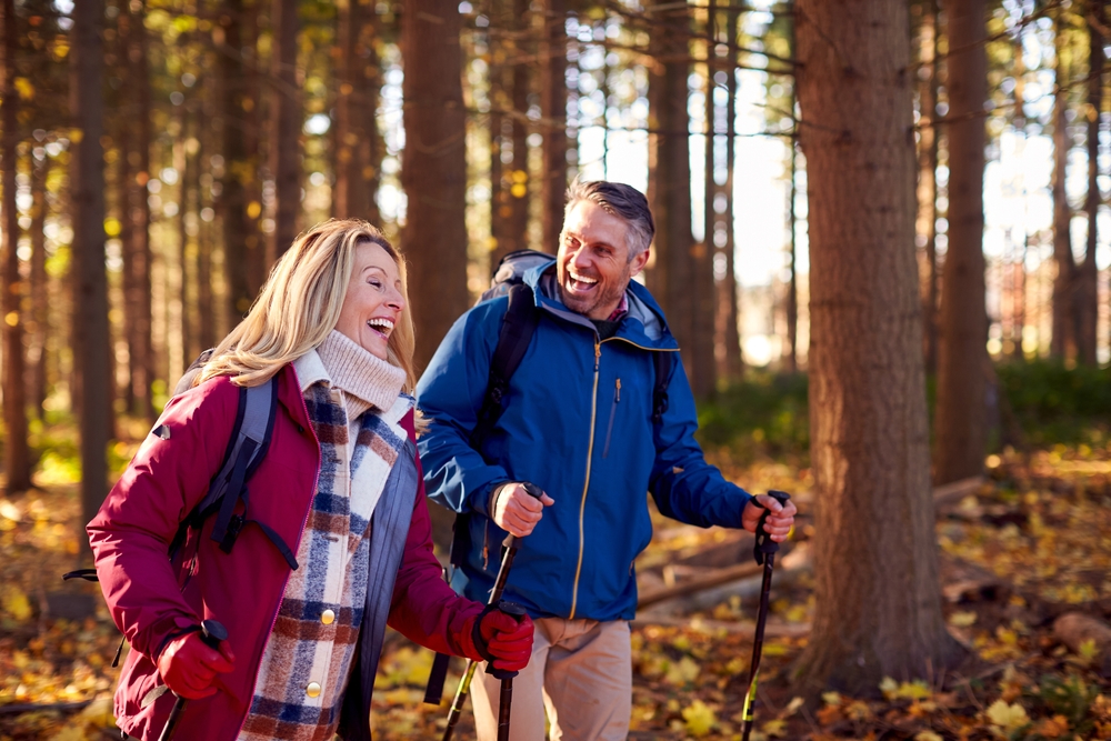 A smiling man and woman wearing jackets and backpacks hike with walking poles through a sunlit forest in autumn, surrounded by tall trees and fallen leaves.