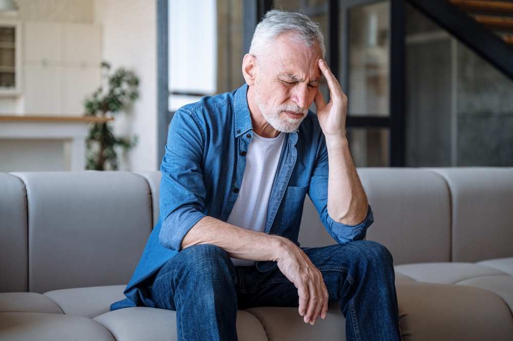 An older man with gray hair sits on a couch, holding his head with one hand and appearing to be in pain or discomfort, possibly experiencing a headache. He is wearing a blue shirt and jeans.