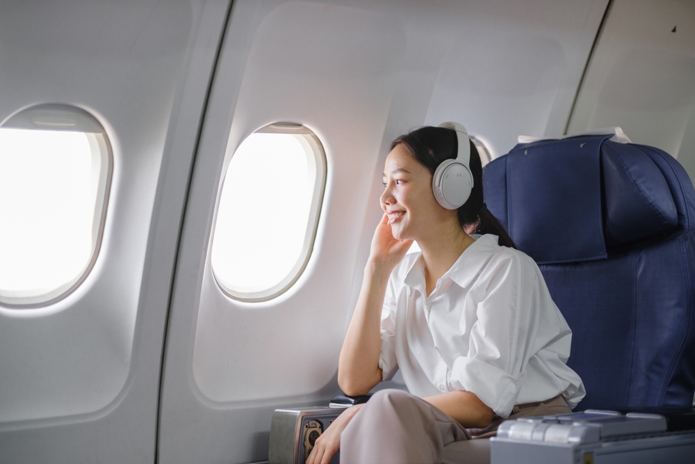 A woman wearing headphones and a white shirt sits in an airplane seat, smiling and looking out the window during her flight.