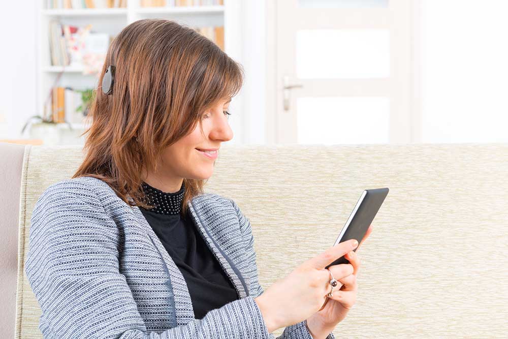 A woman with a cochlear implant sits on a couch, smiling as she looks at a smartphone in her hands. A bookshelf is visible in the background.