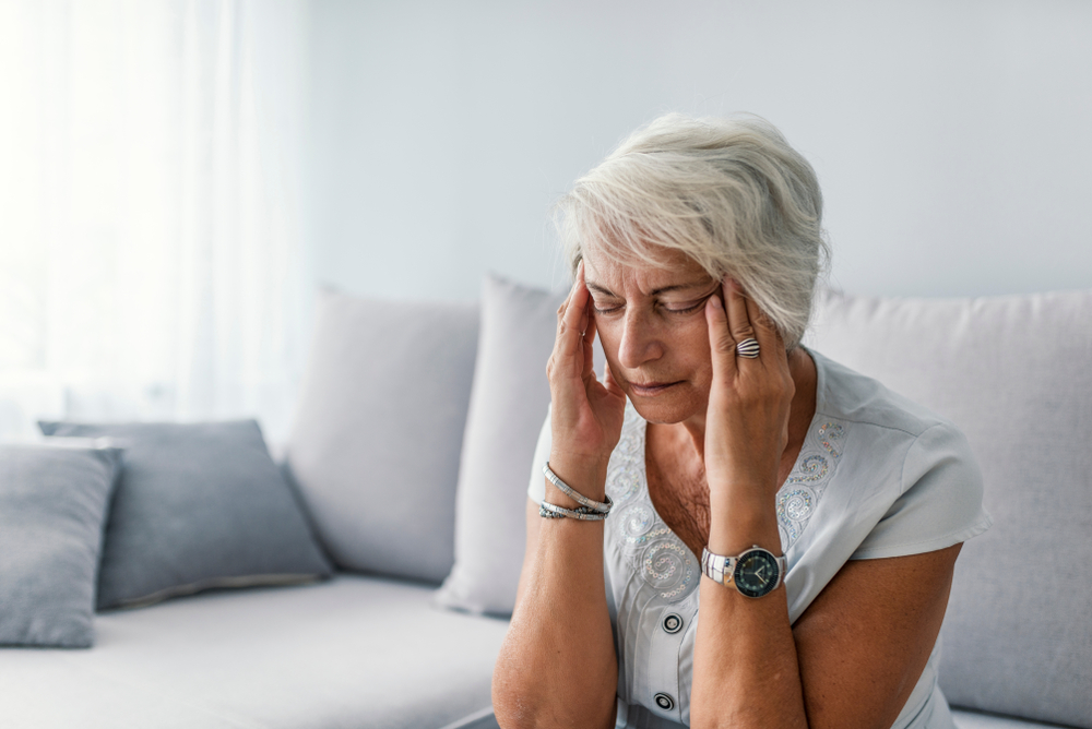 An older woman with gray hair sits on a couch, eyes closed, touching her temples with both hands, appearing to have a headache or feel stressed.