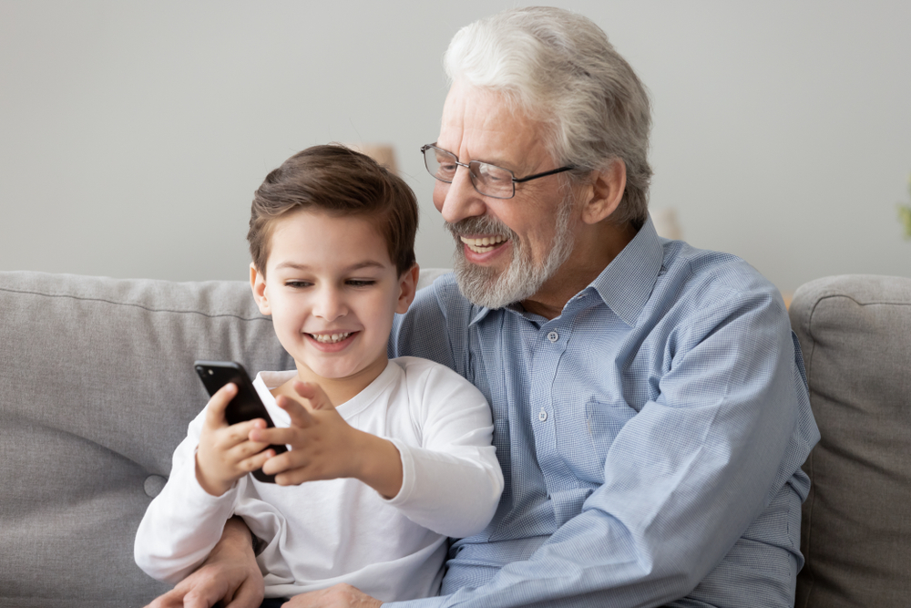 A smiling elderly man with gray hair and glasses sits on a couch, hugging a young boy who is also smiling and holding a smartphone, both looking at the phone screen together.