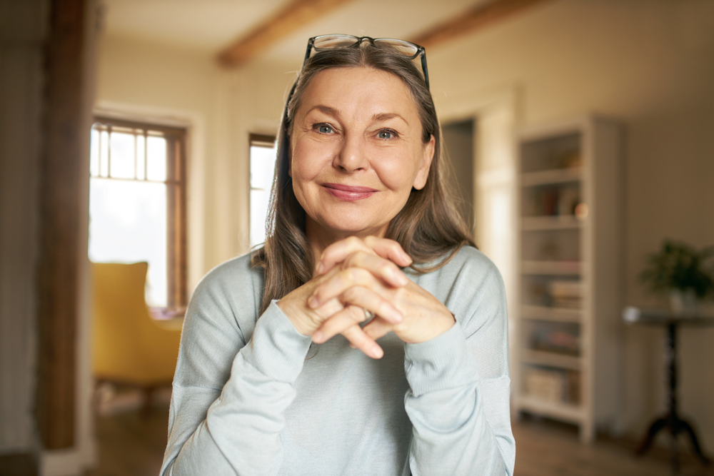 A smiling older woman with long gray hair and glasses resting on her head sits indoors with hands clasped. She wears a light blue top, and the background shows a cozy, sunlit room with shelves and a window.