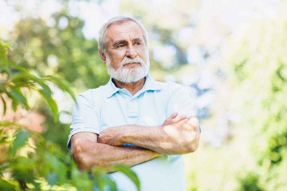 An older man with gray hair and a beard stands outdoors with arms crossed, wearing a light blue polo shirt, looking thoughtful. Lush green foliage and sunlight are visible in the background.
