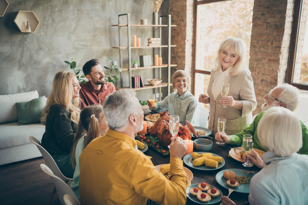 A smiling elderly woman gives a toast at a dining table, surrounded by family members of various ages, enjoying a festive meal together in a cozy, sunlit room.