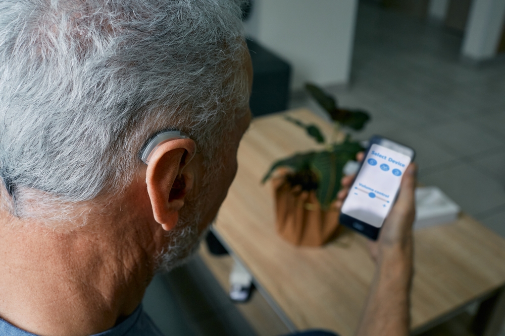 An older man with gray hair wears a hearing aid and holds a smartphone, adjusting hearing aid settings on an app. A potted plant and a table are visible in the background.