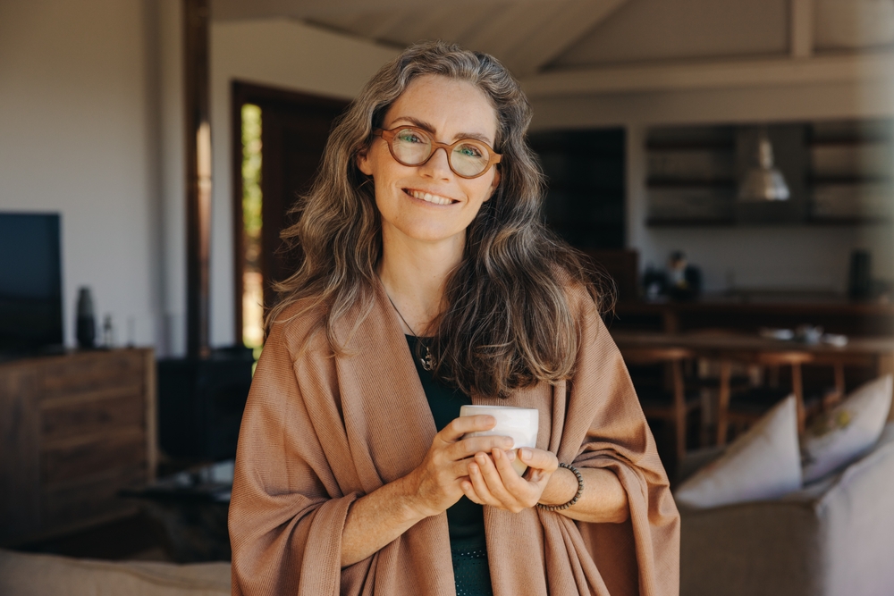 A smiling woman with long gray hair and glasses, wrapped in a brown shawl, stands indoors holding a white mug. The background shows a cozy living room with wooden furniture and shelves.