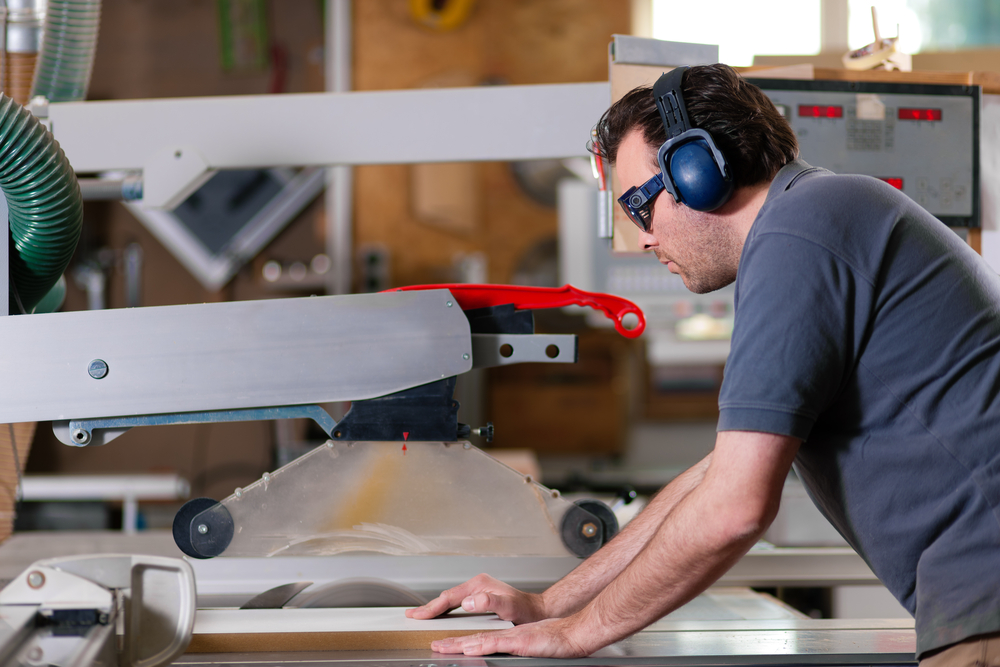 A man wearing safety glasses and ear protection operates a table saw in a woodworking shop, guiding a piece of wood through the blade with both hands.
