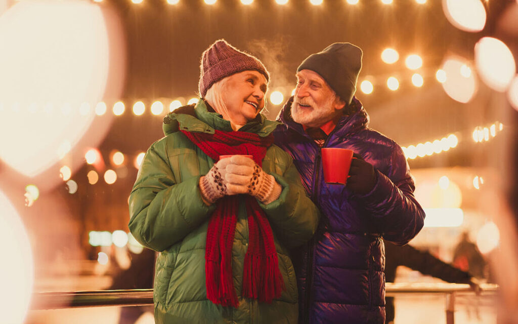 An elderly couple dressed in winter coats and hats smiles warmly at each other outdoors at night, with festive string lights in the background. One person holds a red mug, and both look happy and cozy.