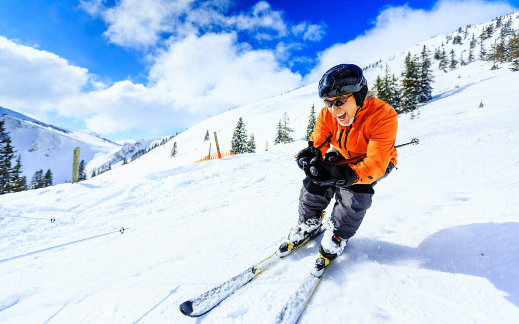 A person wearing an orange jacket, helmet, and goggles skis downhill on a snowy mountain, smiling with trees and a bright blue sky in the background.