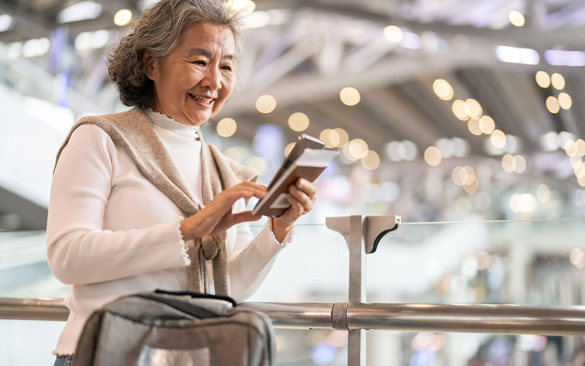 An older woman smiles while holding her passport and phone at an airport. She wears a light sweater draped over her shoulders and stands next to a suitcase, with bright lights and blurred travelers in the background.