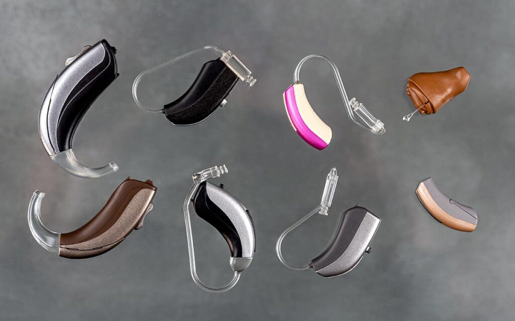 Eight different hearing aids of various shapes, sizes, and colors are arranged on a neutral gray background, showcasing a range of designs and ear molds.