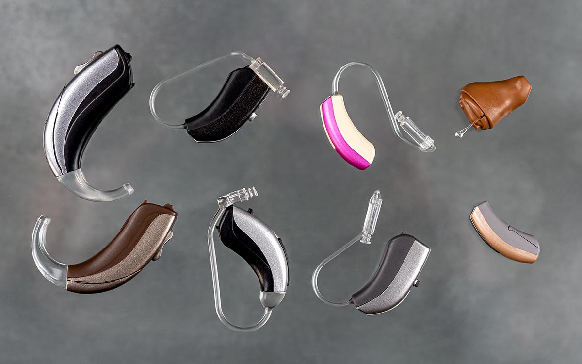 Eight different hearing aids of various shapes, sizes, and colors are arranged on a neutral gray background, showcasing a range of designs and ear molds.