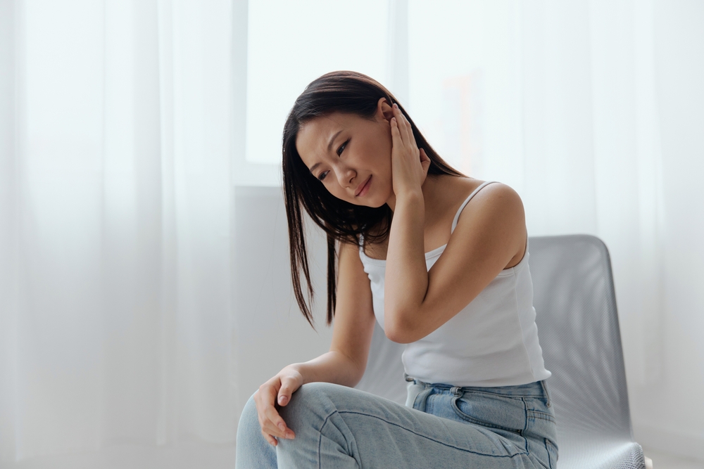 A woman in a white tank top and jeans sits indoors, touching her ear and tilting her head with a pained expression, suggesting she may have ear discomfort or pain.