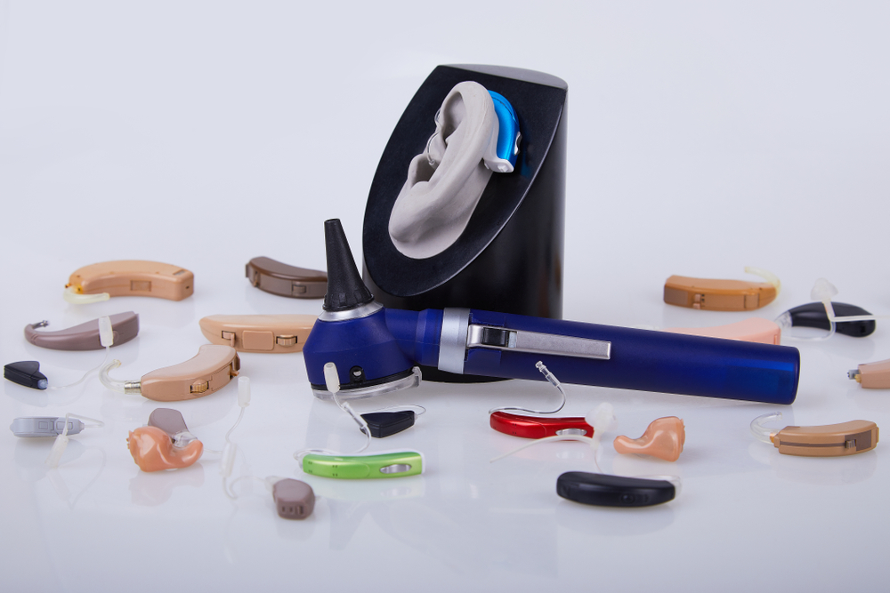 Various hearing aids in different colors and shapes are displayed around an otoscope and a model ear on a stand, all placed on a white background.