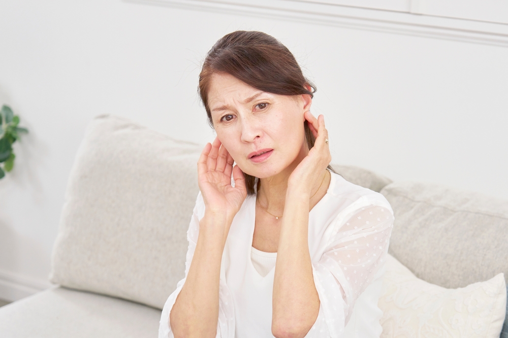 A middle-aged woman sitting on a sofa touches her ears and has a concerned expression, suggesting she may be experiencing ear pain or discomfort.