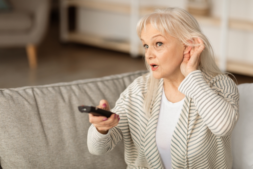 Older woman sitting on a couch, holding a TV remote and cupping her ear as if struggling to hear, with an attentive expression on her face.
