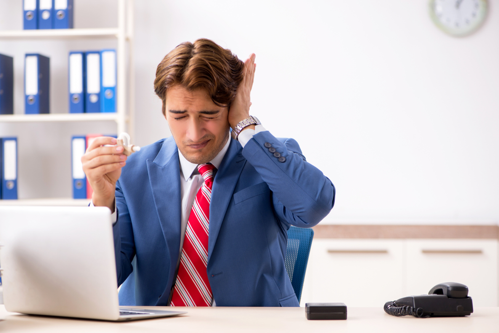 A man in a suit sitting at a desk, grimacing and holding his ear with one hand and a cup in the other, in an office setting with a laptop, phone, and folders in the background.