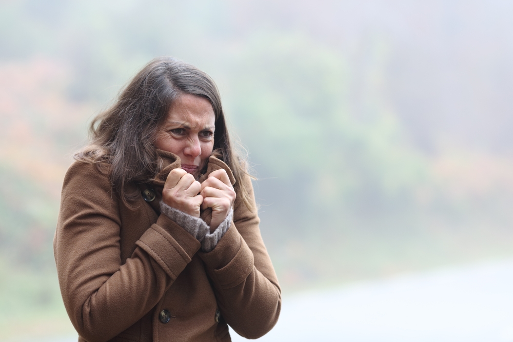 A woman in a brown coat stands outdoors, holding her collar up and shivering from the cold, with a misty, blurred natural background behind her.