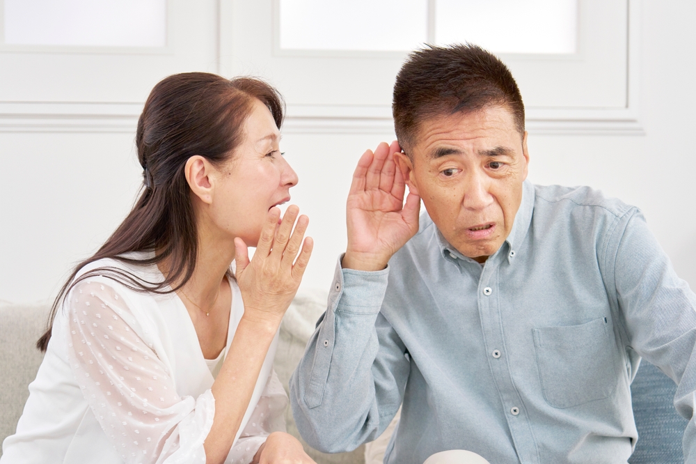 A woman in a white blouse is whispering to a man in a light blue shirt, who is cupping his ear and leaning in to listen, looking confused or concerned. They are sitting indoors on a couch.