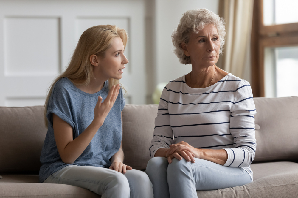 A younger woman sits on a couch speaking emotionally to an older woman in a striped shirt, who looks away with a distant expression and crossed arms. The scene suggests a serious or tense conversation.