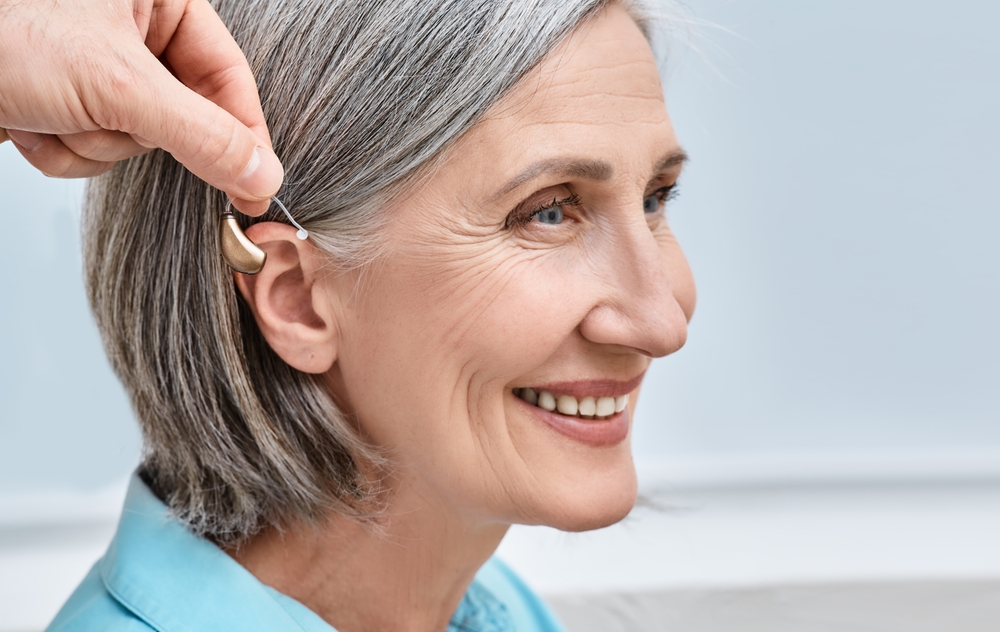 Older woman with short gray hair smiles as someone fits a hearing aid behind her ear, against a light background.