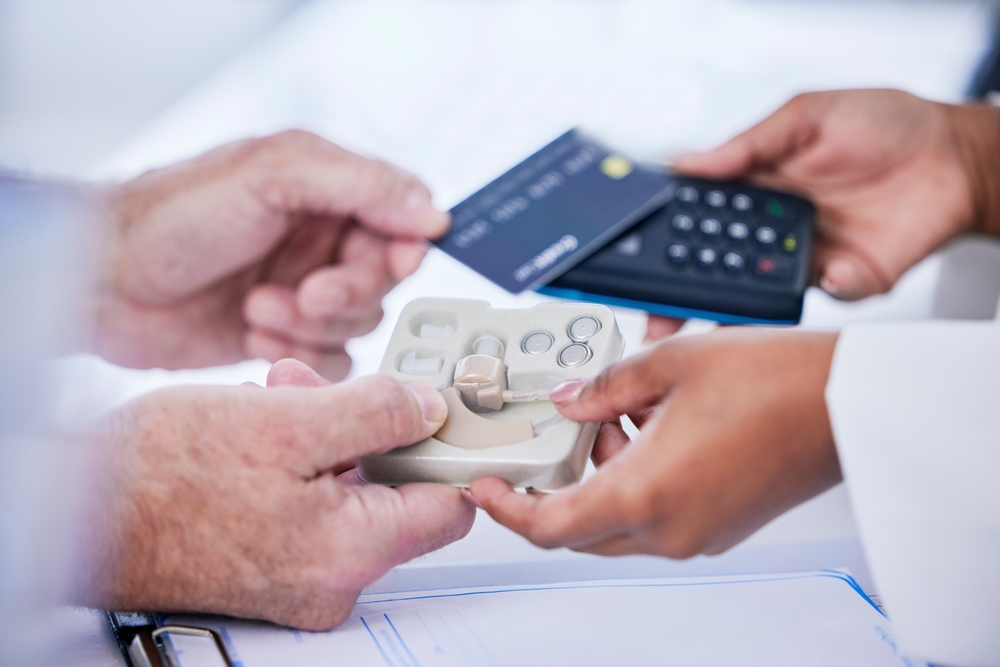 A person holds a credit card and pays with a wireless card reader while another person hands over a tray containing hearing aids and accessories.