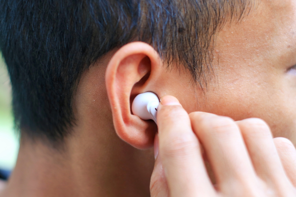 A close-up of a person inserting a white wireless earbud into their ear with their hand. The background is blurred, focusing on the ear and the earbud.