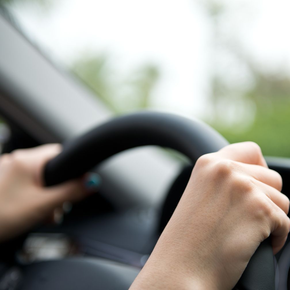 Close-up of a persons hands gripping a black steering wheel while driving a car, with a blurred green background seen through the window.