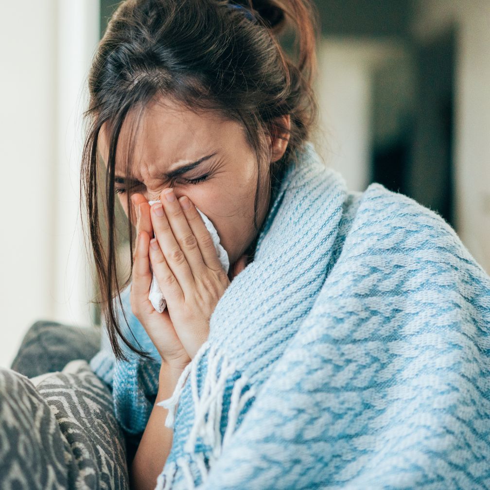 A woman wrapped in a blue blanket sneezes into a tissue, appearing unwell, while sitting on a couch.