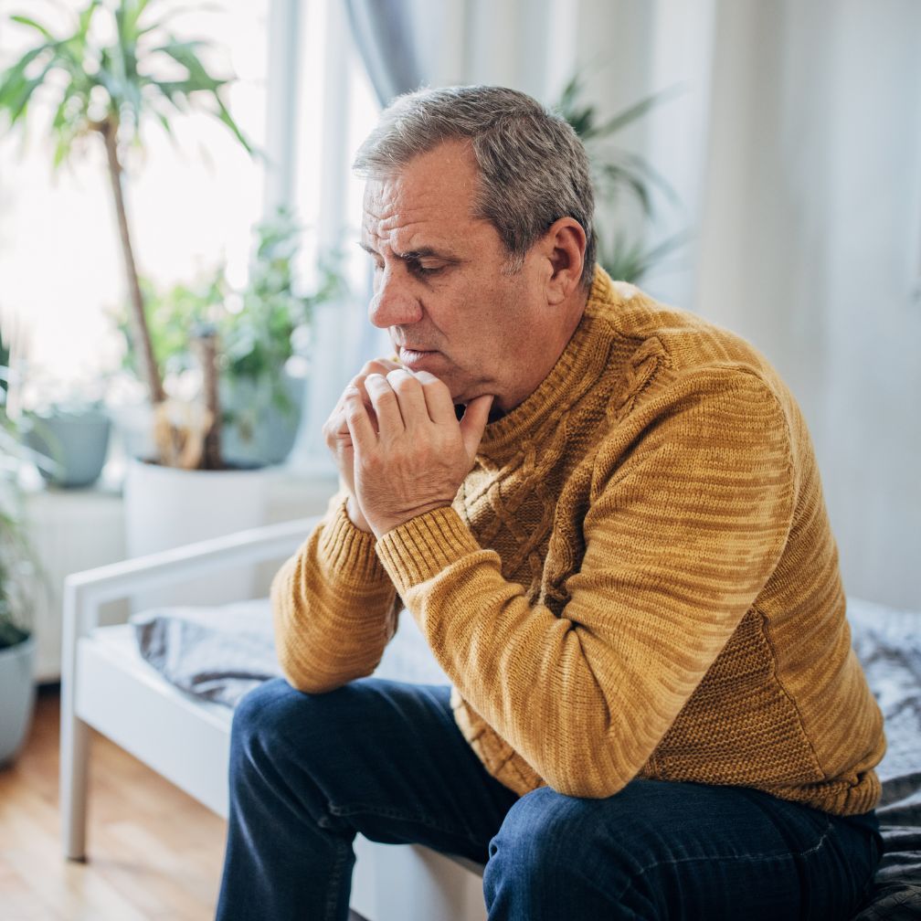 An older man in a mustard sweater sits on the edge of a bed, looking pensive with his hands clasped near his face. There are several plants and soft natural light in the background.