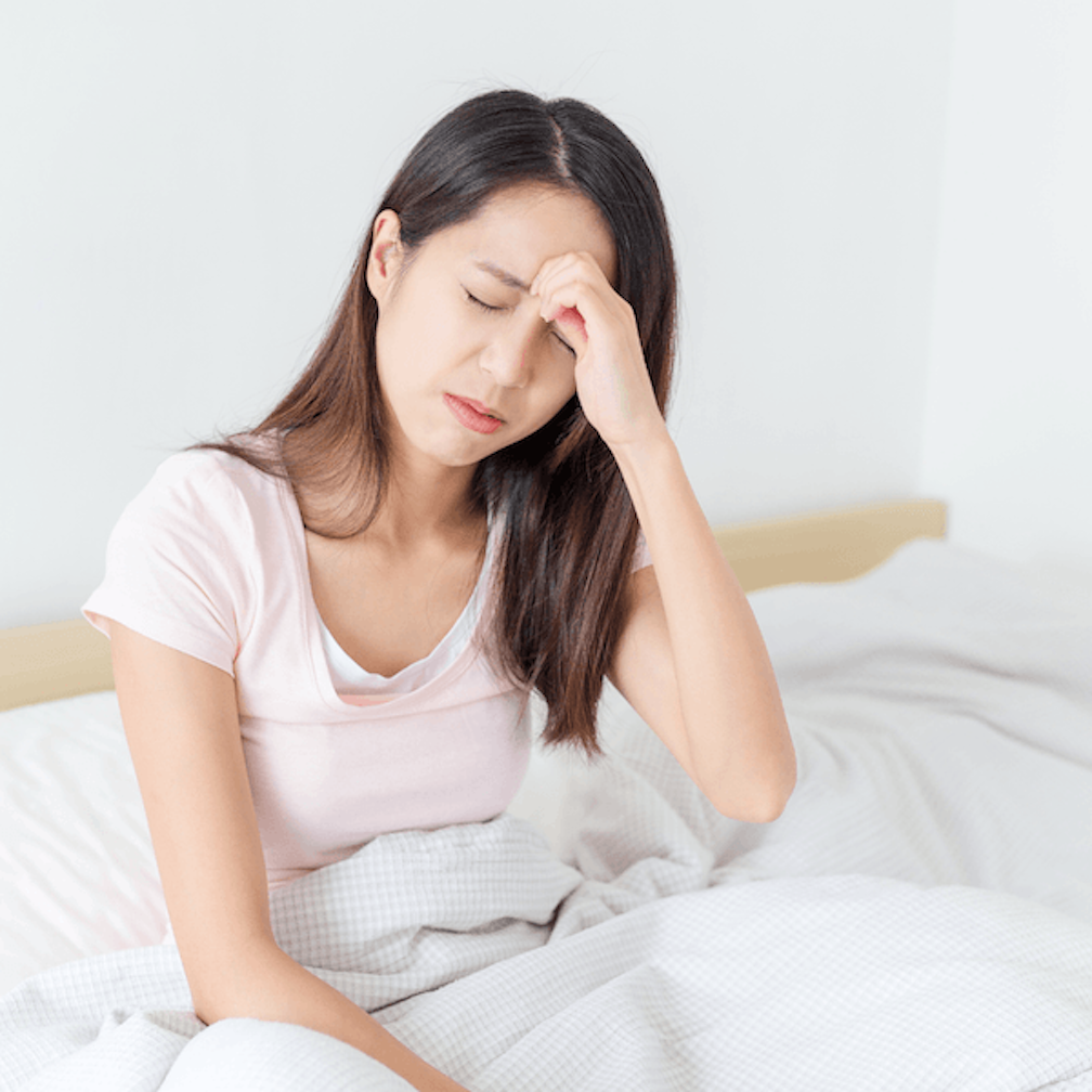 A woman sits on a bed with her eyes closed, holding her forehead and appearing to be in discomfort or pain, possibly due to a headache.