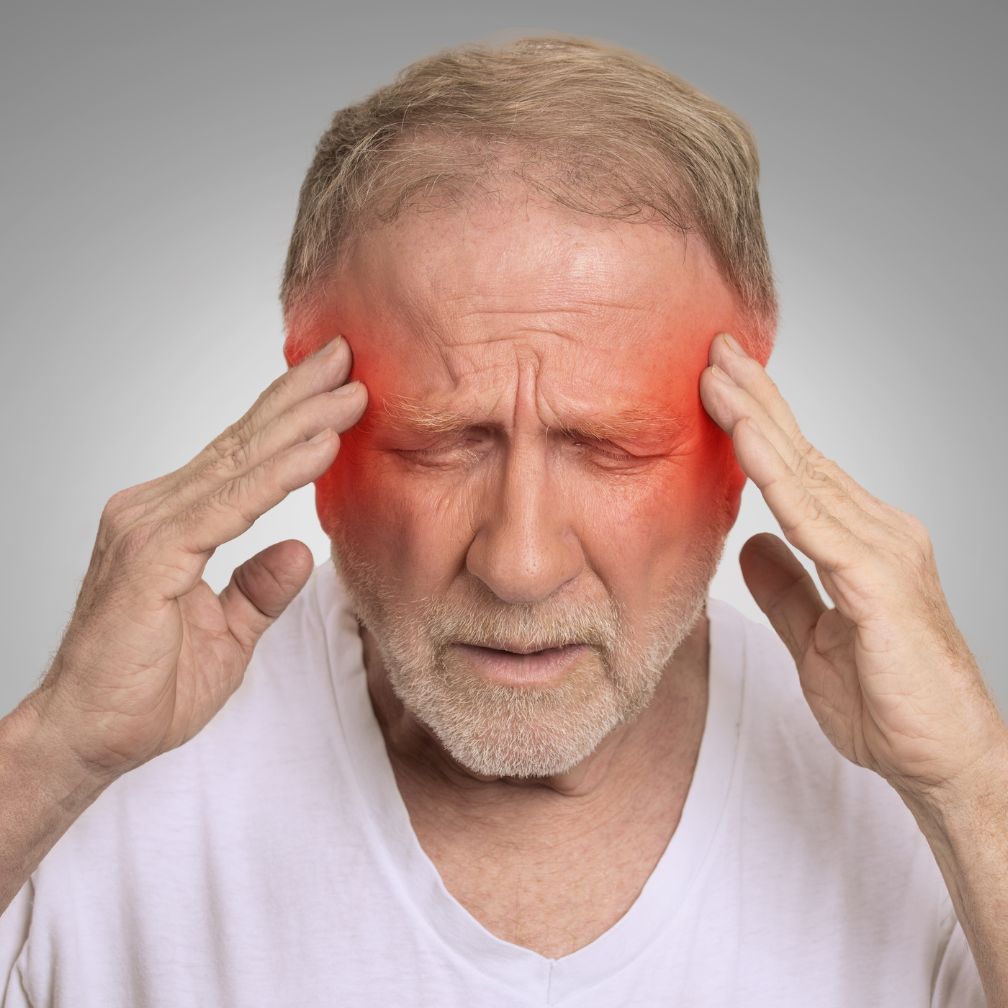 Older man in a white shirt holding his head with both hands, eyes closed, and red highlights around his temples, indicating he is experiencing a headache or migraine.