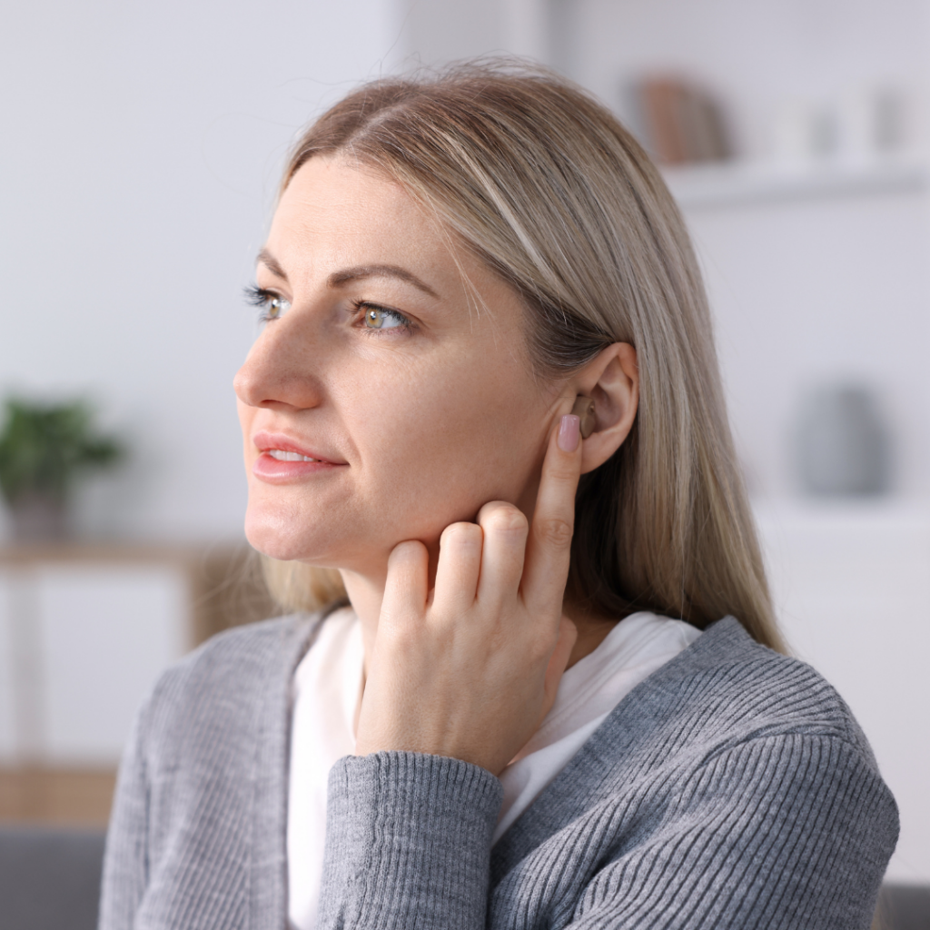 A woman with blonde hair touches her ear with her finger, possibly indicating discomfort or listening closely. She wears a gray sweater and is indoors with blurred shelves and a plant in the background.