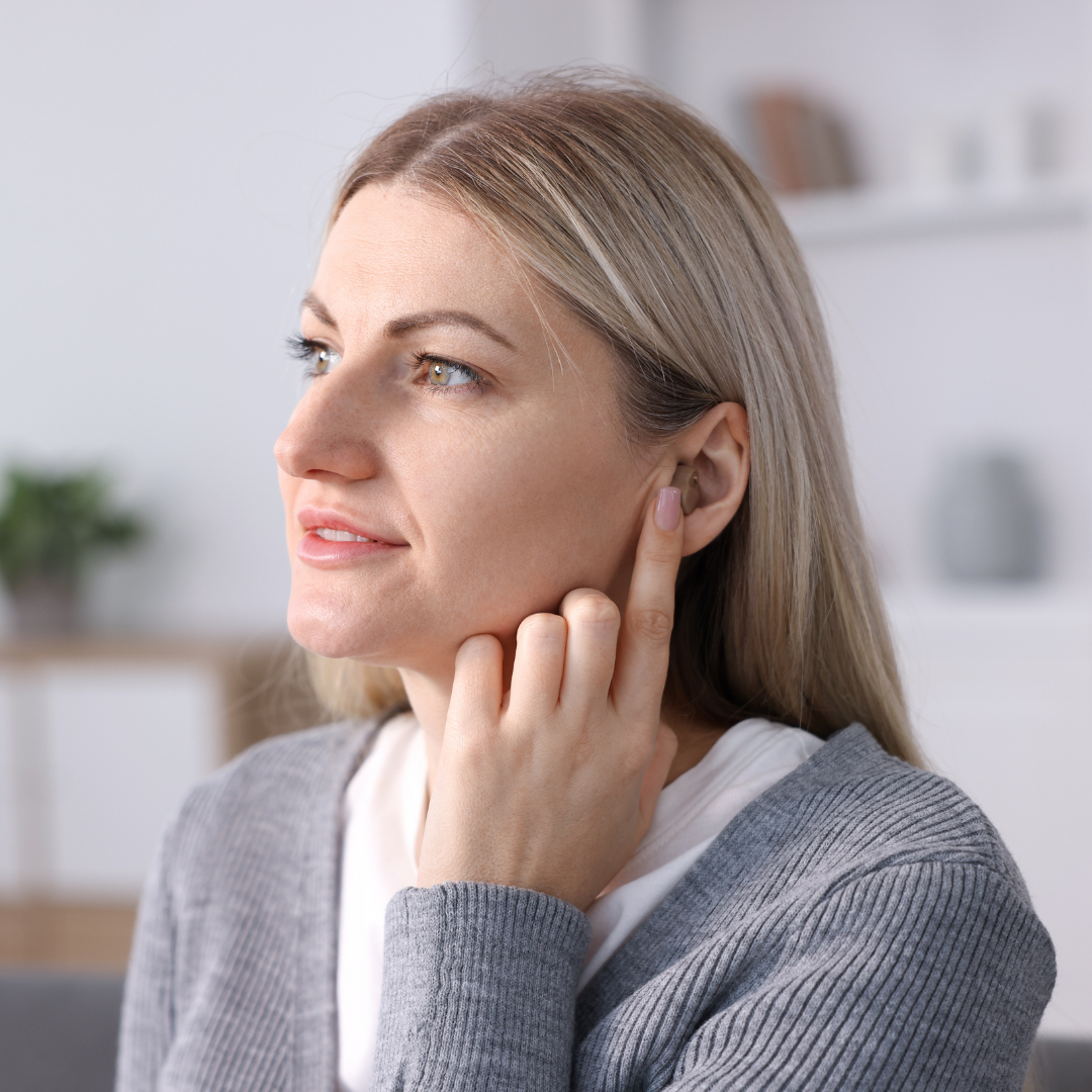 A woman with blonde hair touches her ear with her finger, possibly indicating discomfort or listening closely. She wears a gray sweater and is indoors with blurred shelves and a plant in the background.